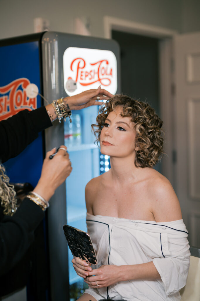 Makeup artist puts finishing touches on brides makeup in front of a vintage Pepsi Cola vending machine at The Ballroom at Ellis Preserve