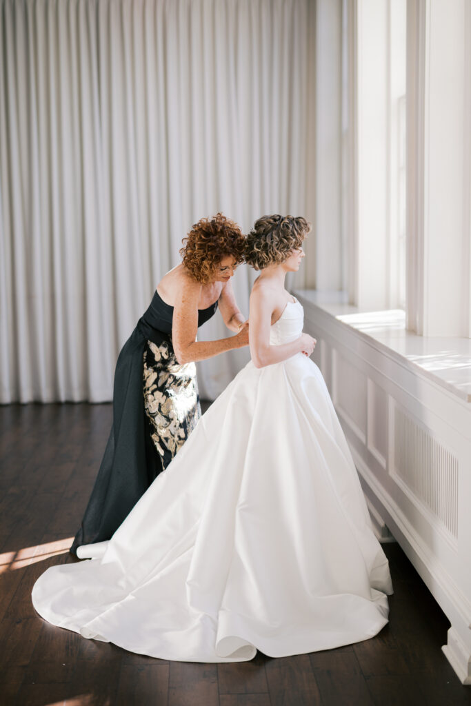 Mother of the bride buttons the back of brides ballgown in front of elegant white draping and large windows at The Ballroom at Ellis Preserve bridal suite to get ready for a Villanova church wedding