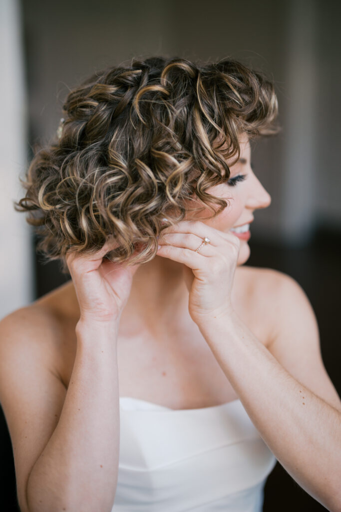 Bride with curled crown braid hairstyle puts an earring in her ear as she gets ready for her Villanova church wedding ceremony