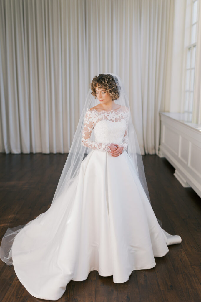 Bride wears an elegant white ballgown wedding dress with a long sleeved lace top overlay and chapel length veil
