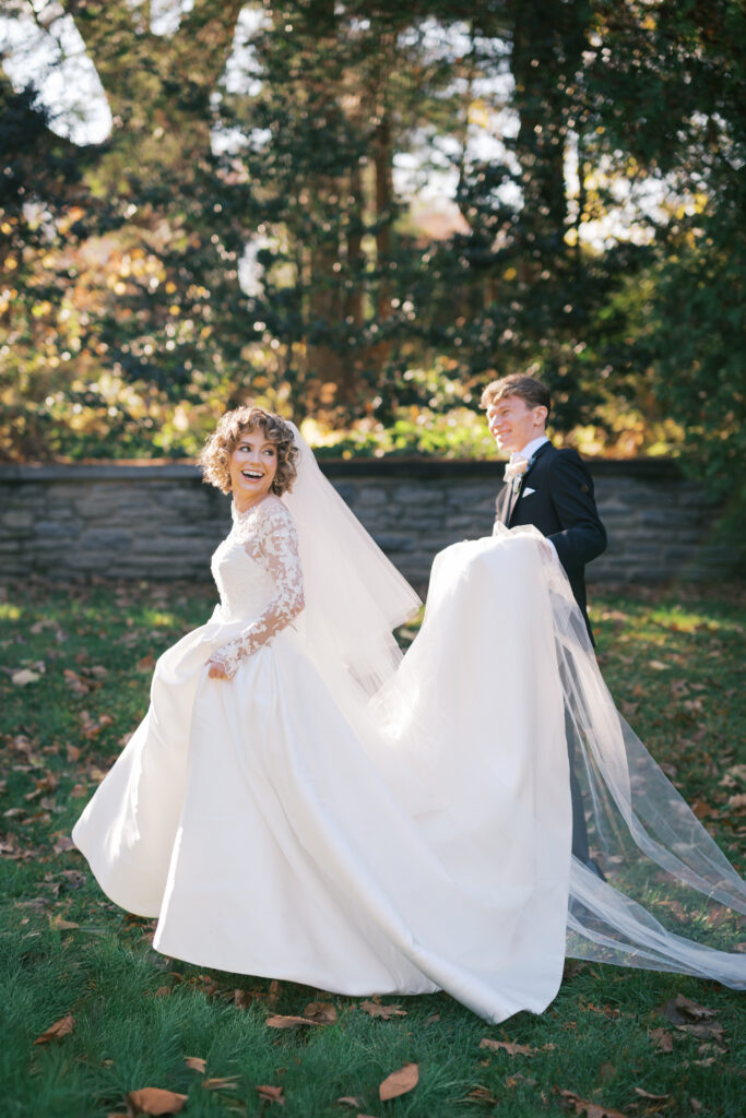 Groom carries bride's ballgown dress train through the Stoneleigh Gardens as they pose for their first look portraits in Villanova PA