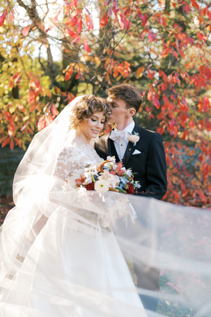 Bride and groom stand in front of vibrant red and gold fall foliage as brides veil softly flutters around them in the breeze at Stoneleigh Gardens