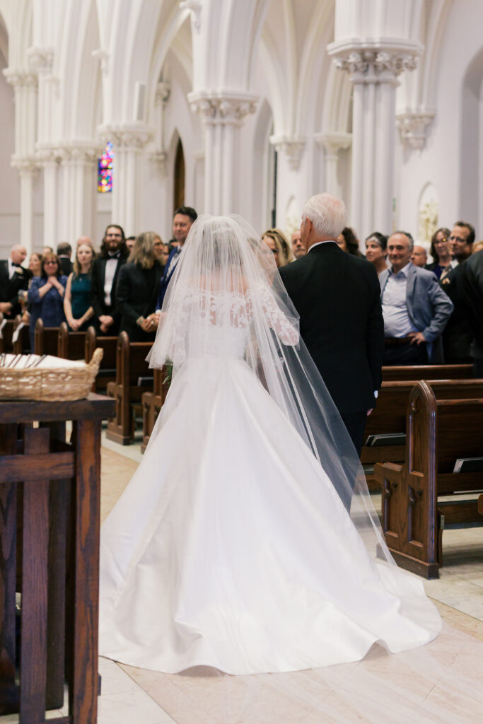Bride and her father get ready to walk down the aisle inside of St. Thomas of Villanova Church as guests look on from wooden pews