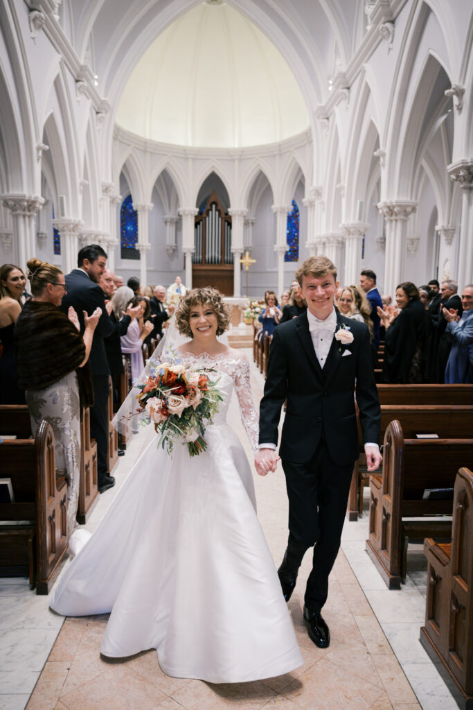 Bride and groom hold hands and smile as they walk back down the aisle after their St. Thomas of Villanova church ceremony concludes. Soaring Gothic arches frame them in the background