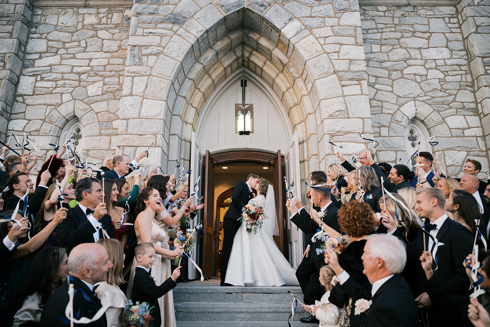 Bride and groom kiss on the front steps of Villanova Church while their guest surround them on either side while smiling, cheering, and waving ribbon streamers