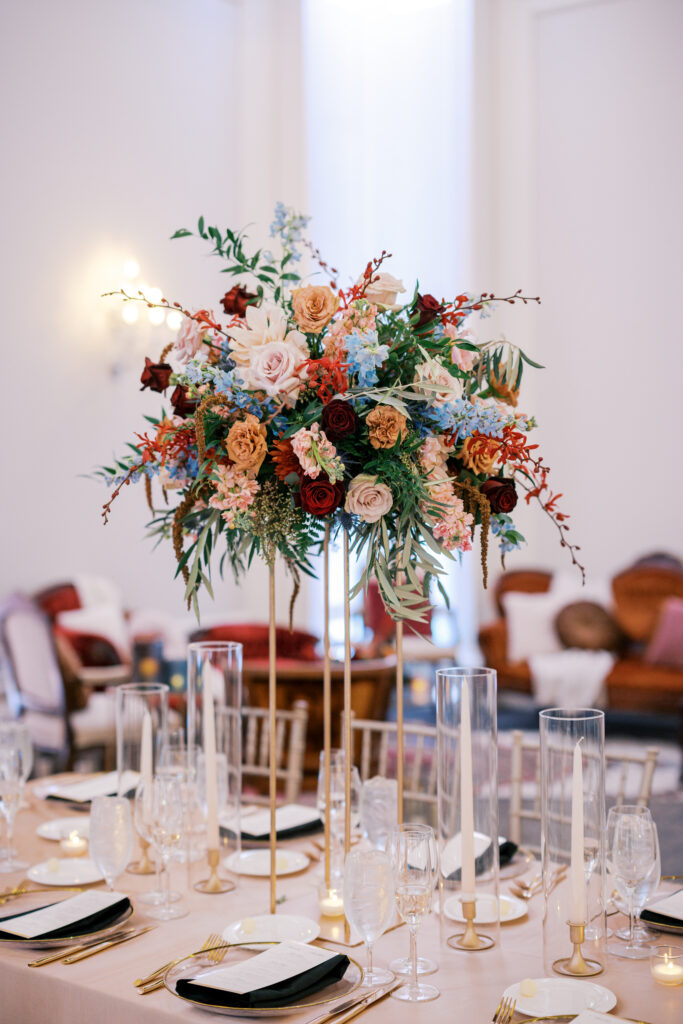 Elegant tablescape features black and white dishware and linens with a large fall floral centerpiece accented by blue, blush, and deep burgundy flowers and greenery