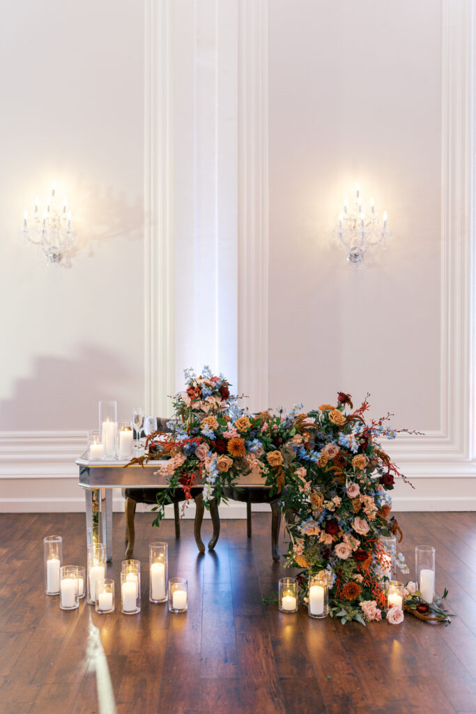 Sweetheart table is decorated with an oversized arrangement of trailing florals in blush, blue, and burgundy and surrounded by romantic votive candles at the Ballroom at Ellis Preserve