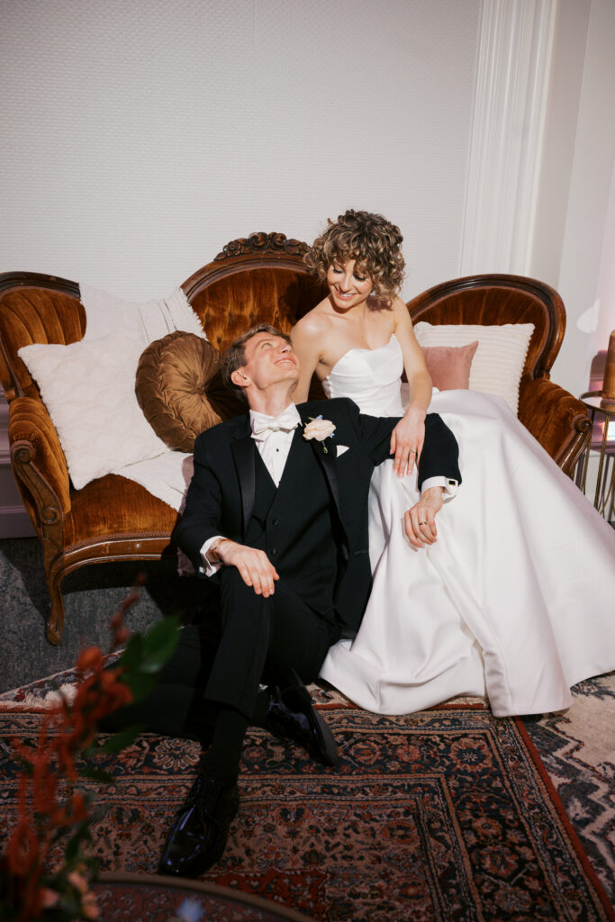 Vintage style photo of a bride and groom lounging on a rust colored velvet couch at The Ballroom at Ellis Preserve