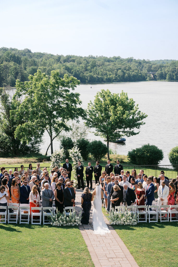 Romantic lakefront wedding ceremony in progress with bride walking down aisle white florals Lake House Inn Perkasie PA