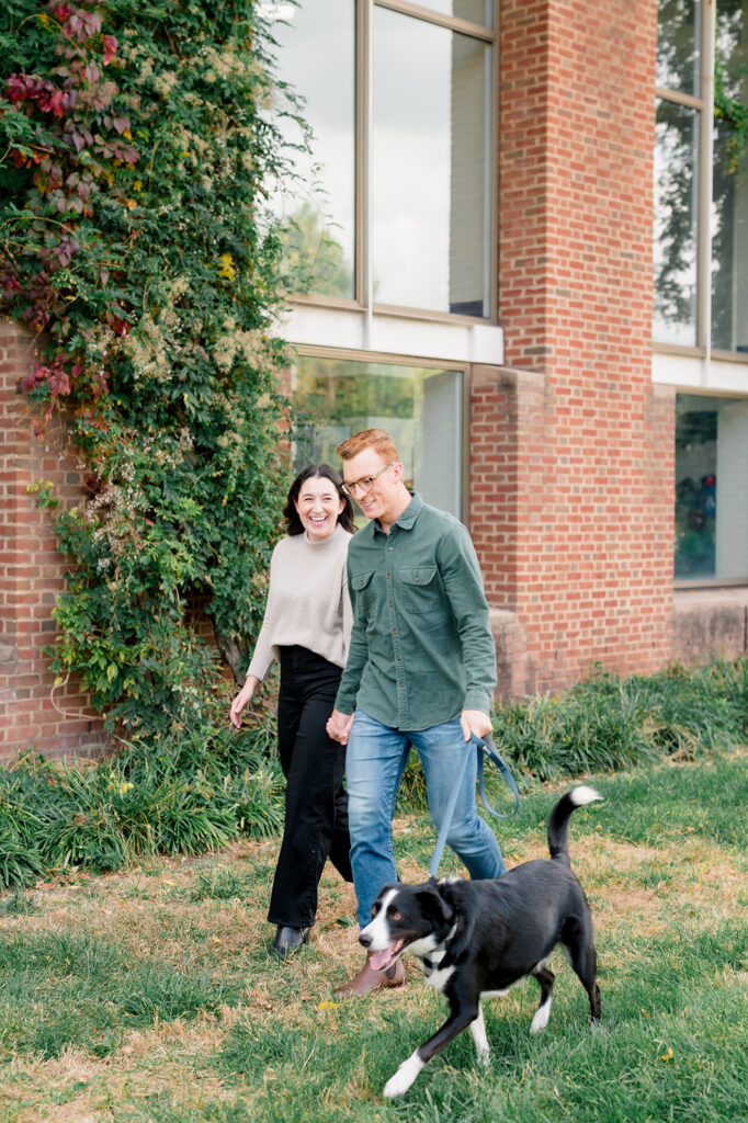 Couple walks their black and white dog on a leash in front of an ivy covered brick building in Old City Philly during their engagement session