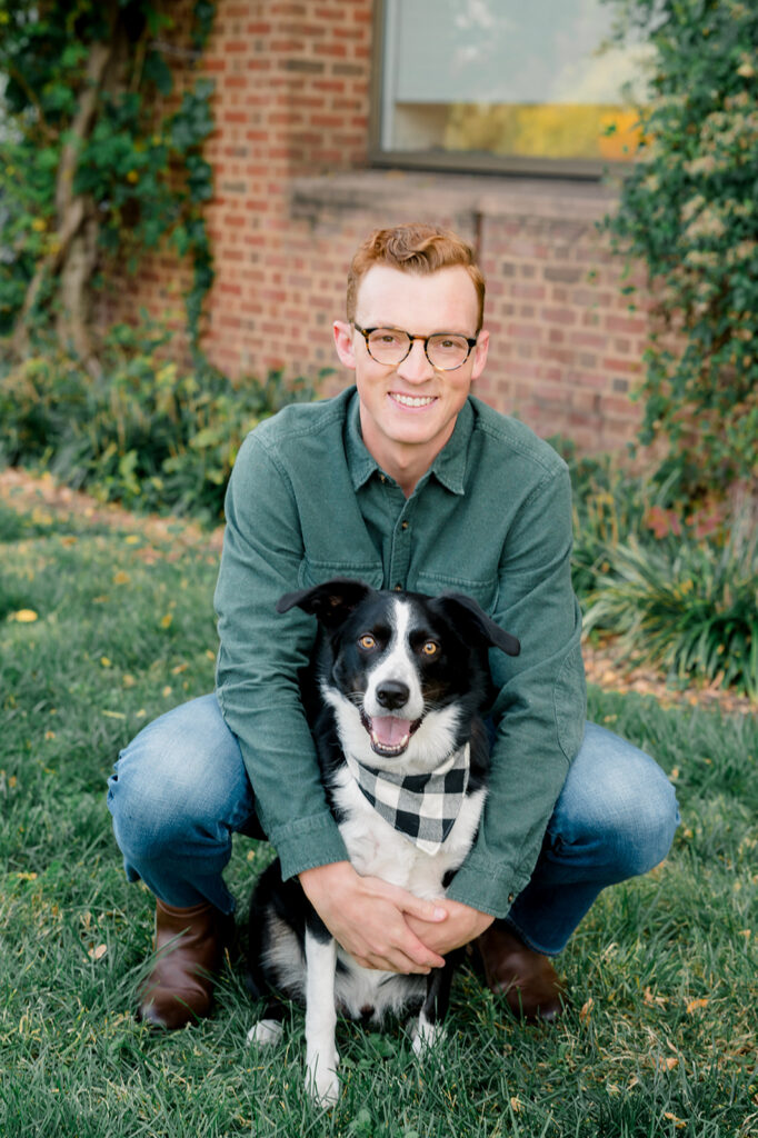 Red haired man wearing glasses and a green button down hugs his black and white dog wearing a black and white checked bandana