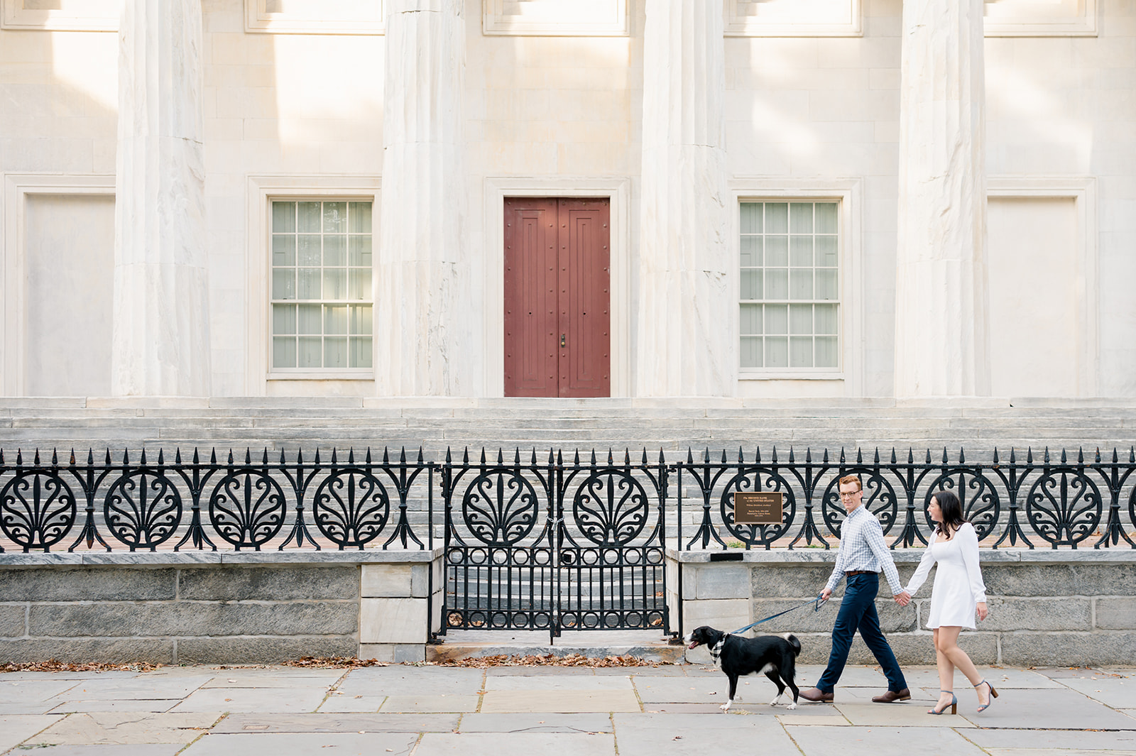Couple walks along a sidewalk in front of an ornate wrought iron fence and stone column building in Old City Philly. Their black dog is on a leash walking in front of them.
