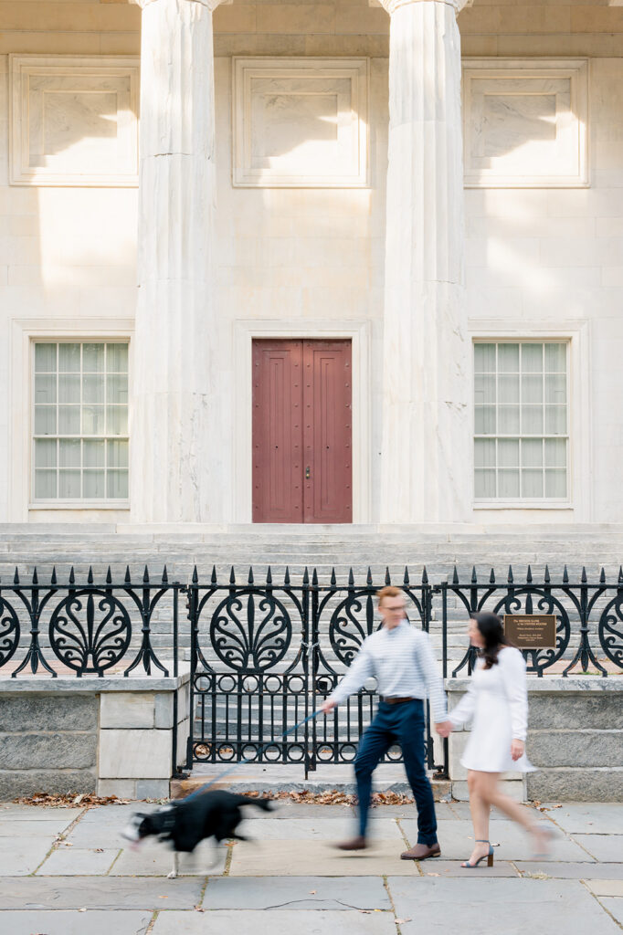 Couple walks along a sidewalk in front of an ornate wrought iron fence and stone column building in Old City Philly. Their black dog is on a leash and their bodies are blurred