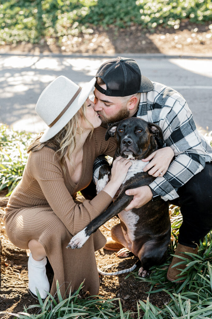 Engaged couple wearing hats and neutral colors hugs their black and white dog between them as they kiss during their engagement photos as Longwood Gardens.