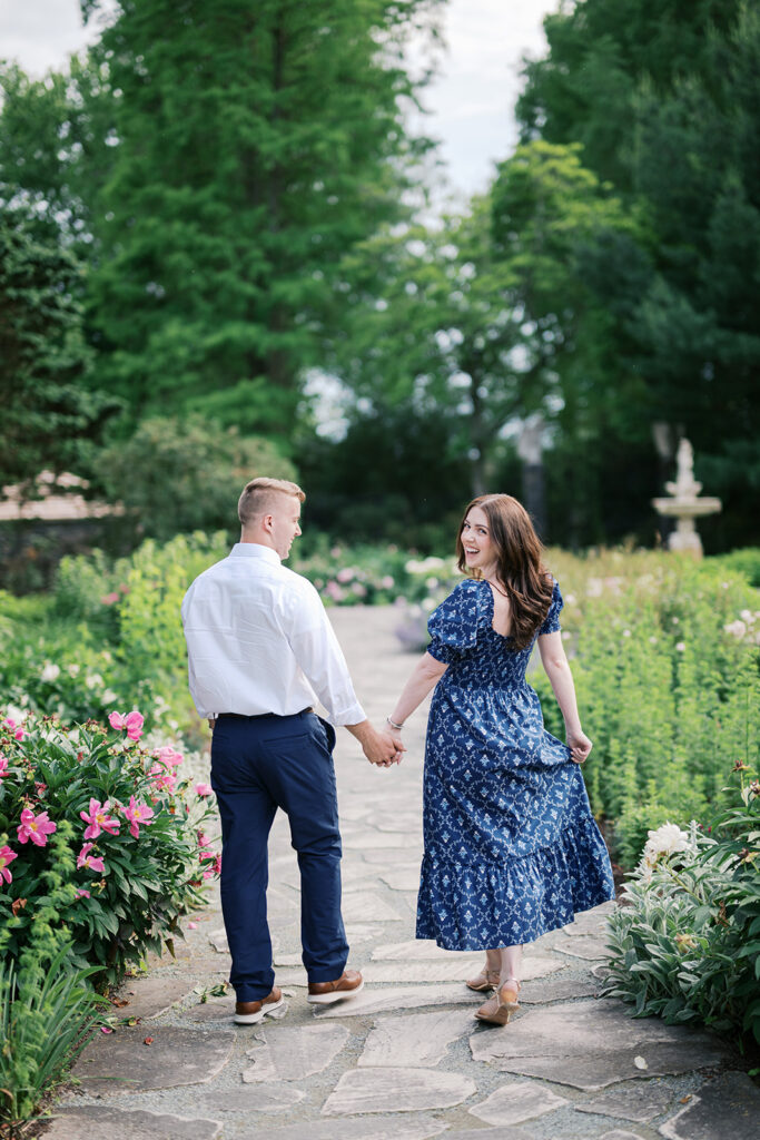 Couple walking hand in hand along stone path lined with pink roses during their engagement photos at Marian Coffin Gardens