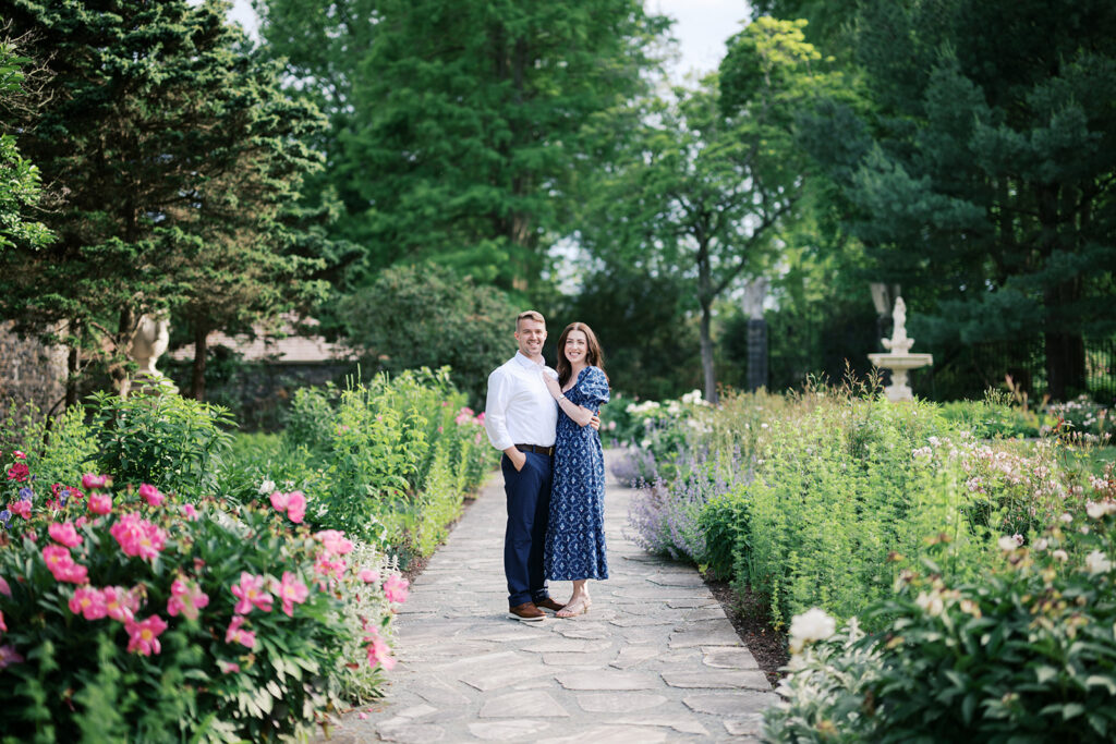 Engaged couple smiling together on the stone garden path surrounded by blooming pink roses and lavender during engagement photos at Marian Coffin Gardens