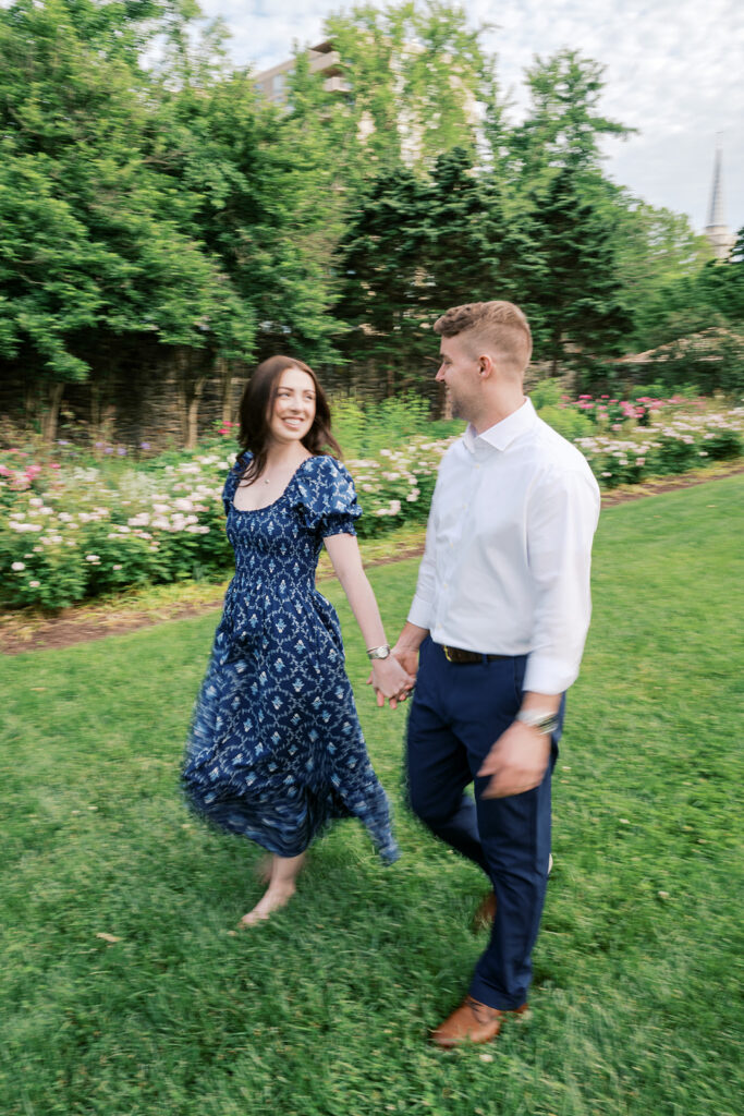 Couple laughing and running together on the green lawn during candid engagement session at Marian Coffin Gardens in DE