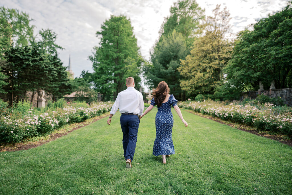 Couple walking hand in hand away from camera along a wide grass lawn bordered by roses during an engagement photoshoot