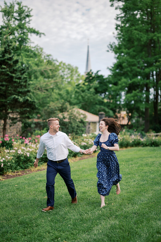 Joyful couple running on the lawn with pink roses in the background at Marian Coffin Gardens engagement session