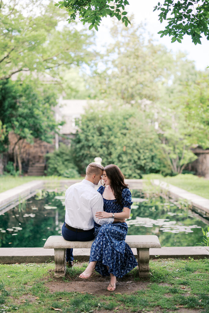 Engaged couple sitting together on a stone bench beside the lily pond at Marian Coffin Gardens engagement session in New Jersey