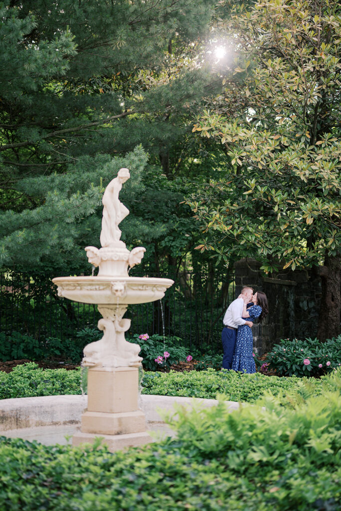 Couple kissing behind an ornate tiered garden fountain with sun flare during their engagement photoshoot at Marian Coffin Gardens