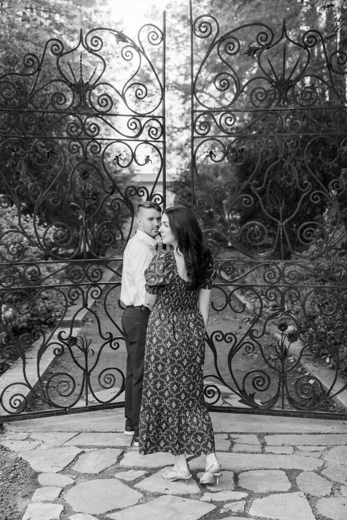 Black and white photo of man leading a woman through ornate wrought iron garden gates during their engagement photoshoot at Marian Coffin Gardens