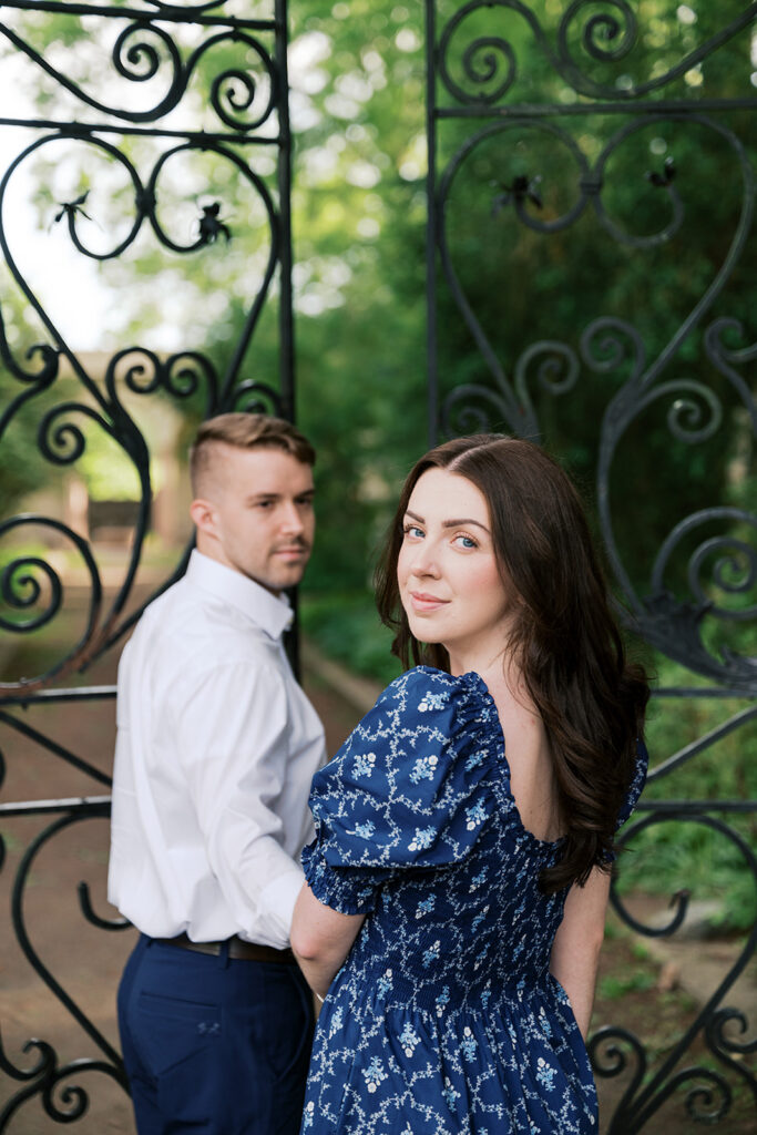Engaged couple posing at decorative wrought iron garden gates during engagement session at Marian Coffin Gardens NJ