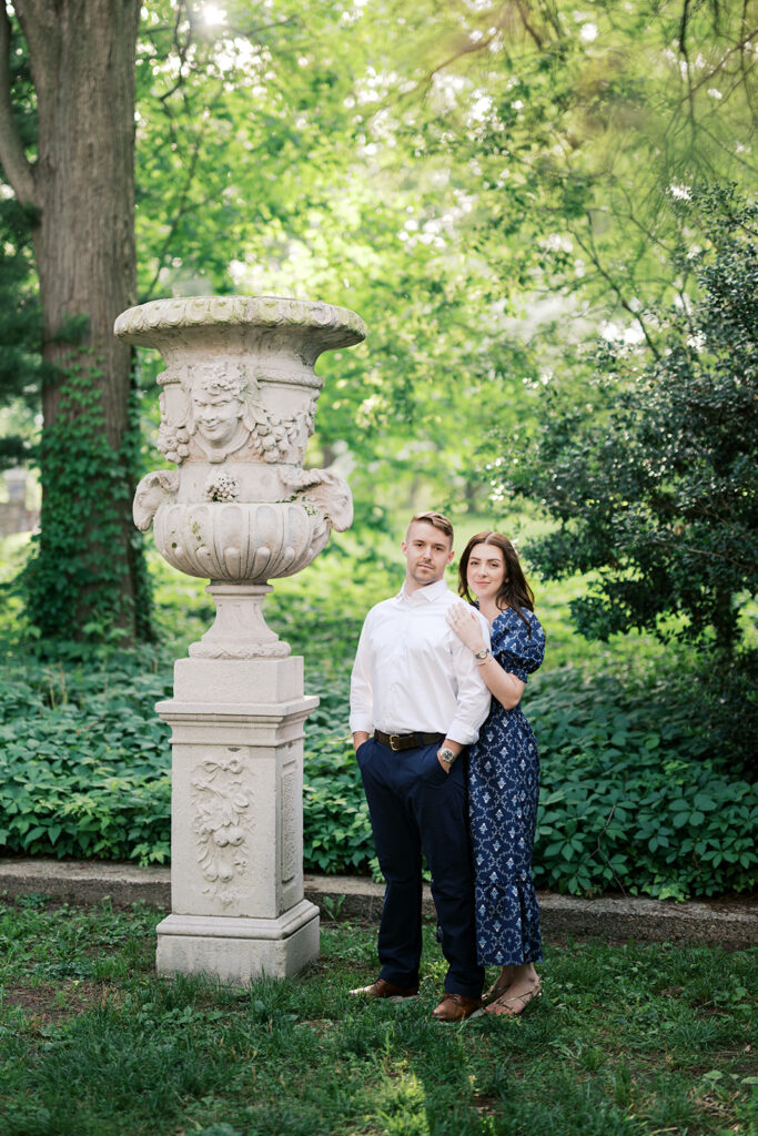 Couple standing beside a large carved stone urn pedestal in lush greenery during Marian Coffin Gardens engagement session