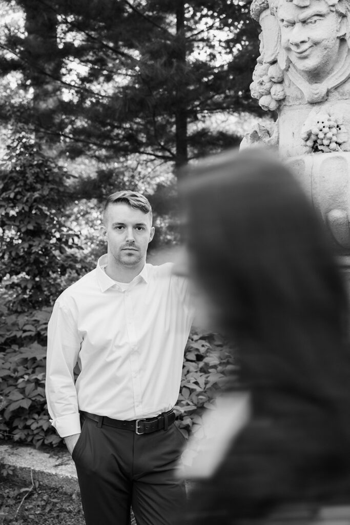 Black and white editorial portrait of groom leaning against carved stone urn with blurred foreground