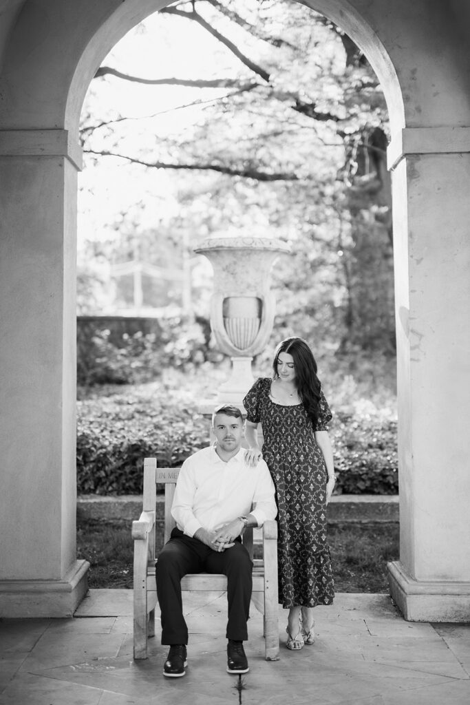 Black and white photo of couple posed under stone arch colonnade with garden urn during engagement session at Marian Coffin Gardens