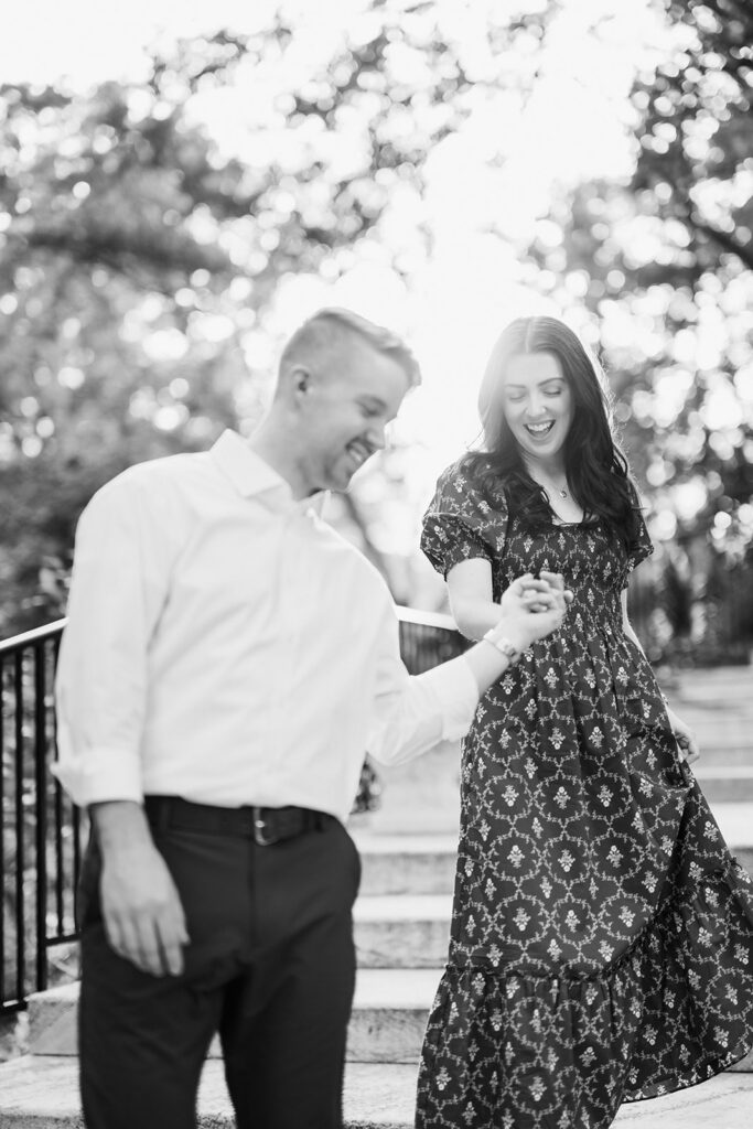Black and white candid photo of laughing couple holding hands in backlit golden light during their engagement session