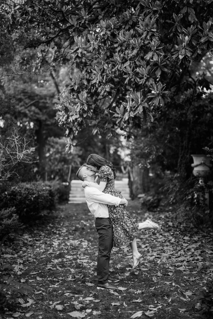 Black and white romantic photo of groom lifting bride-to-be for a kiss along a magnolia-lined garden path during engagement session at Marian Coffin Gardens