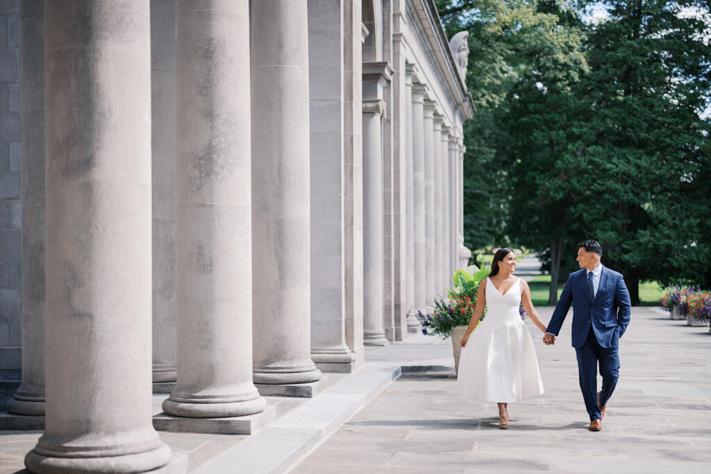Couples photoshoot engagement couple walking along Nemours Estate stone colonnade in white midi dress and navy suit Wilmington Delaware summer