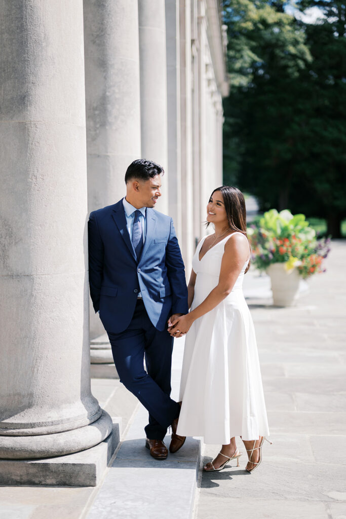 Engagement photo aesthetic couple by stone column at Nemours Estate colonnade white midi dress with gold strappy heels and navy suit Delaware