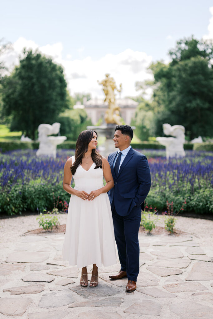 Romantic couples photography couple in white midi dress and navy suit at Nemours Estate formal garden with gold Neptune fountain and purple salvia Wilmington Delaware