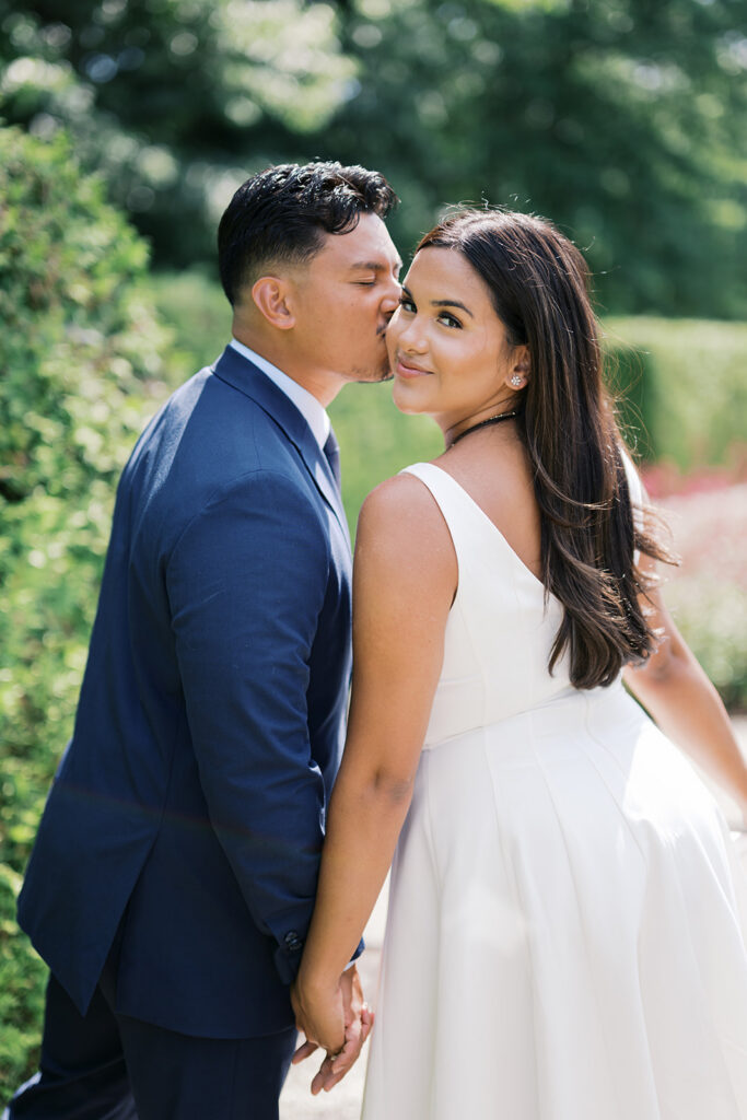 Engagement photo poses woman in white sleeveless midi dress looks at camera while fiancé kisses her cheek at Nemours Estate engagement session