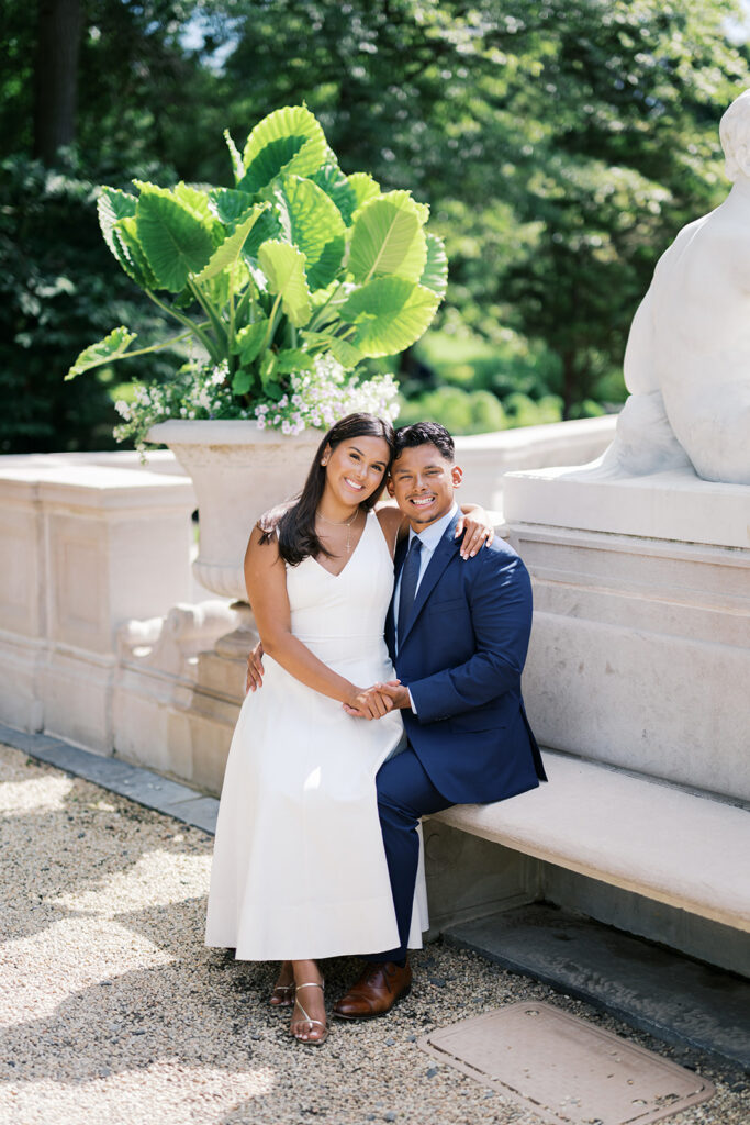Nemours Estate engagement photography couple seated on stone bench in white midi dress and navy suit summer engagement photo outfit ideas
