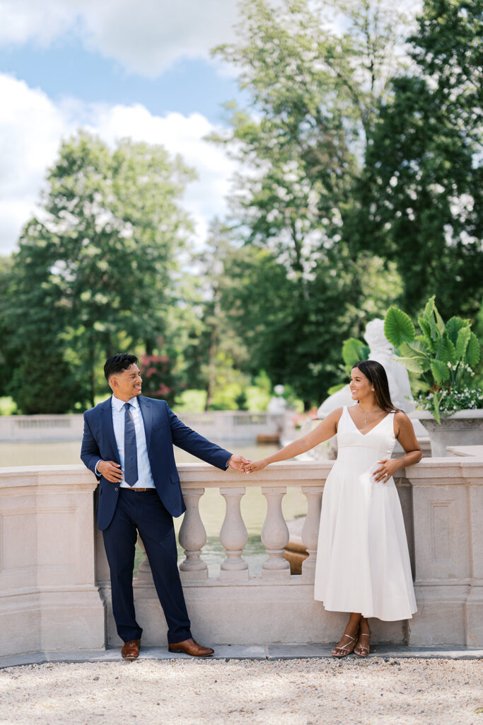 Nemours Estate engagement photos couple holding hands across stone balustrade terrace in white dress and navy suit Wilmington Delaware