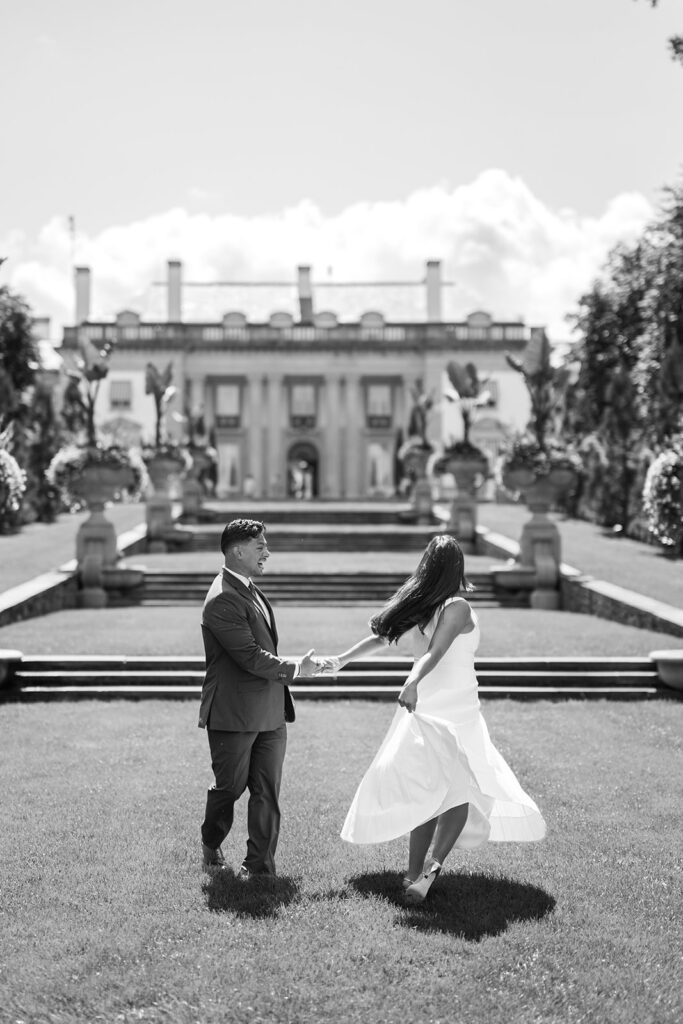Black and white engagement photos couple twirling on Nemours Estate lawn with Nemours mansion facade and classical statues in background Delaware