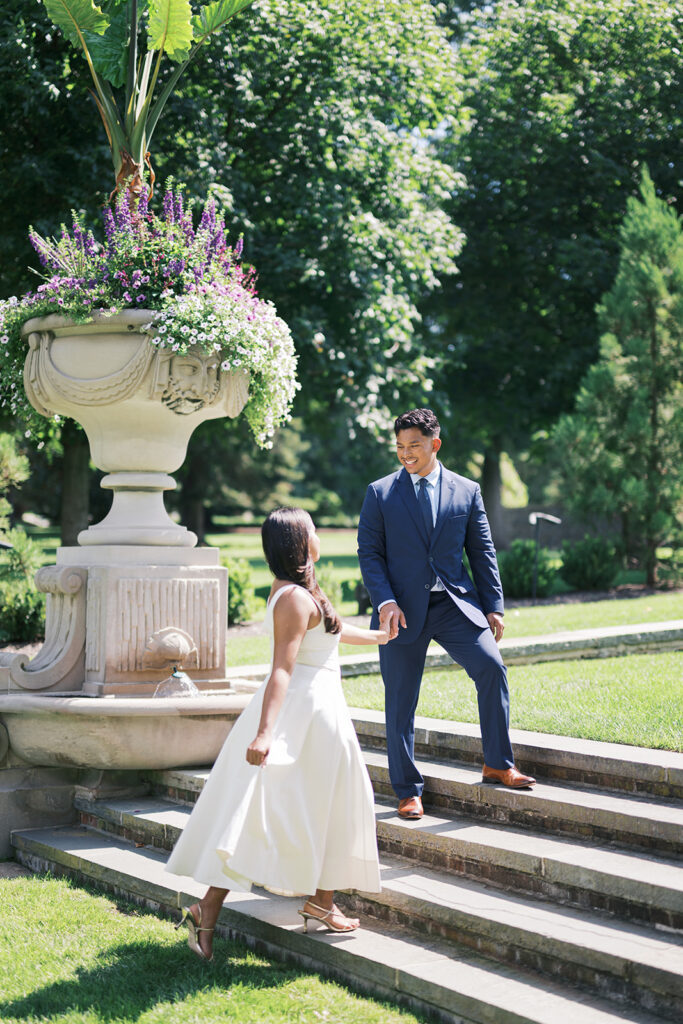 Couple walks up Nemours Estate stone garden steps Wilmington Delaware in engagement photo outfit ideas white midi dress with gold strappy heels and navy suit