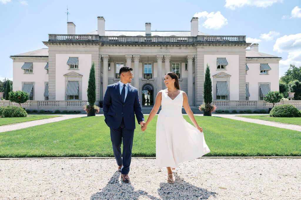 Engagement photo inspiration couple walking hand in hand laughing in white midi dress and navy suit on gravel path at Nemours Estate Delaware