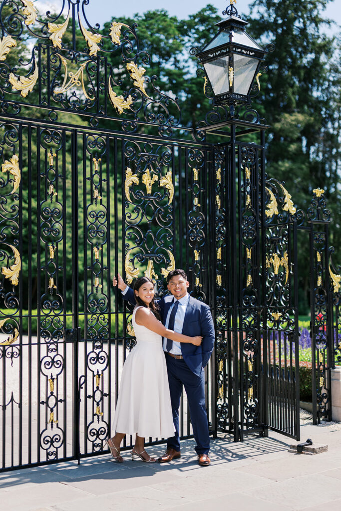 Nemours Estate engagement photography couple at ornate black and gold iron gate in white midi dress and navy suit Wilmington Delaware summer