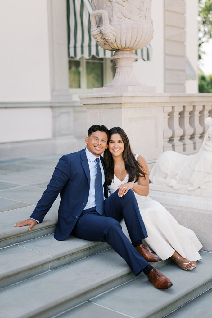 Couples photoshoot engagement seated portrait on Nemours mansion portico steps white v-neck midi dress and navy suit with engagement ring detail Delaware