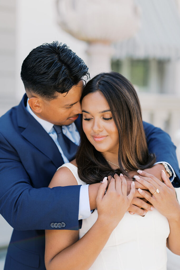 Engagement photo aesthetic close portrait man embraces woman from behind with engagement ring detail white sleeveless dress navy suit Nemours Estate Wilmington Delaware