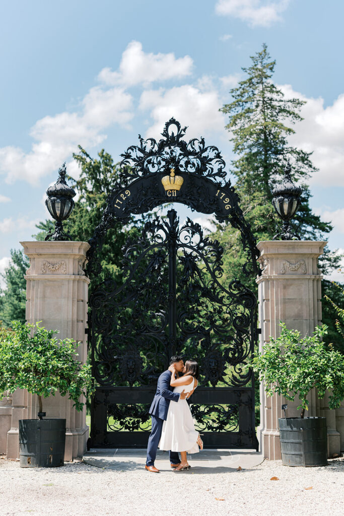 Romantic couples photography couple kissing under ornate iron crown gate at Nemours Estate in white midi dress and navy suit Wilmington Delaware