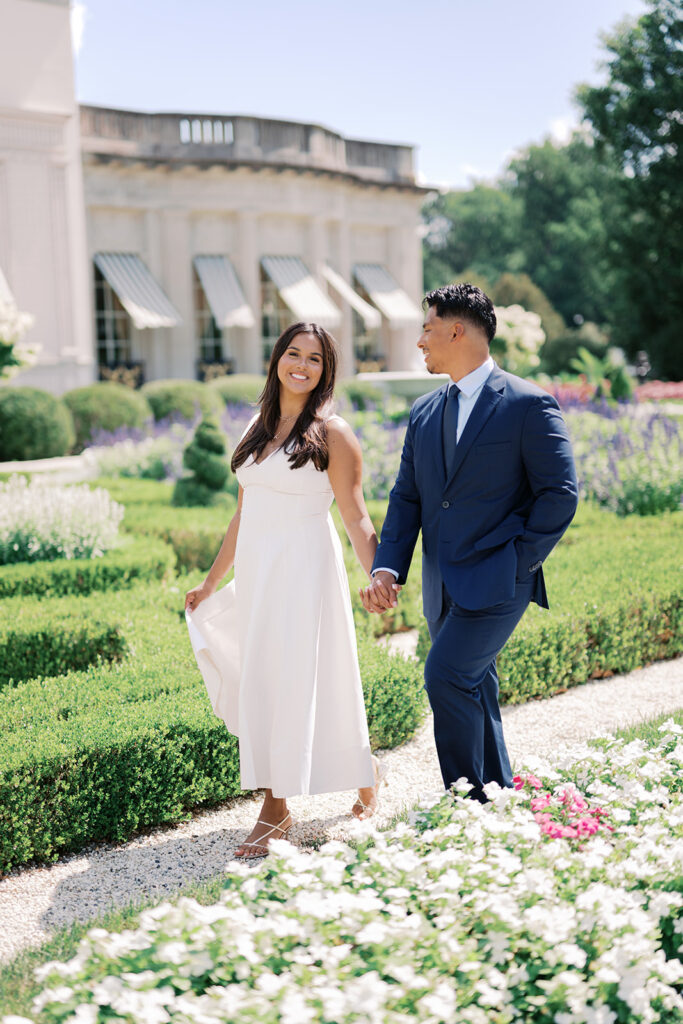 Engagement photo outfits summer white midi dress with strappy sandals and navy suit walking through Nemours Estate formal parterre garden Wilmington Delaware