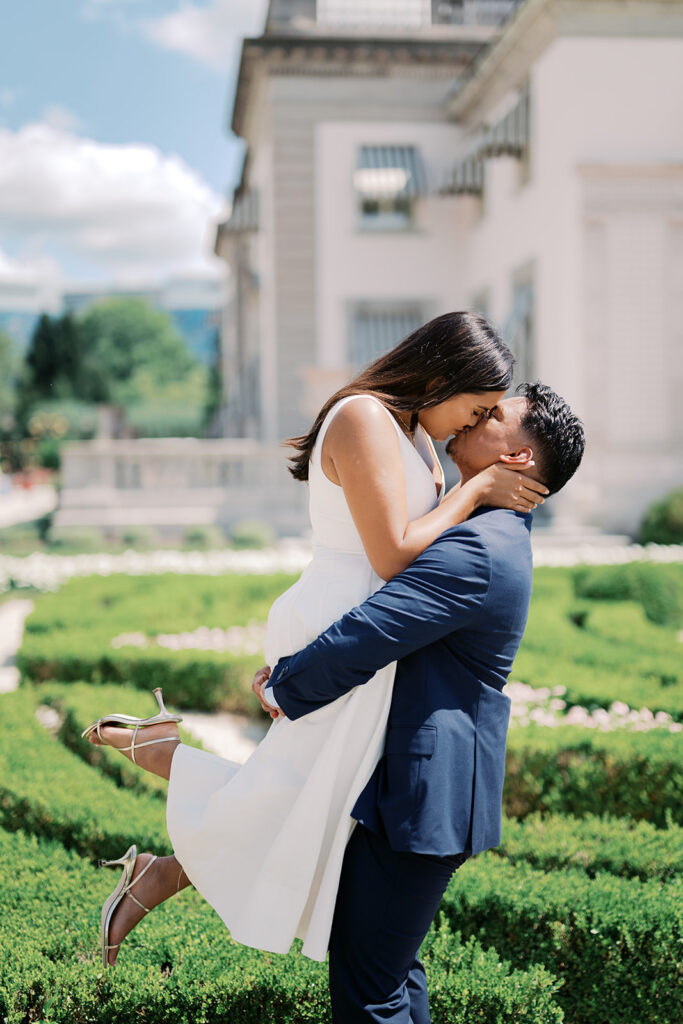 Couples photoshoot poses man lifting woman in white dress with gold strappy heels in Nemours Estate formal garden Wilmington Delaware summer engagement photos