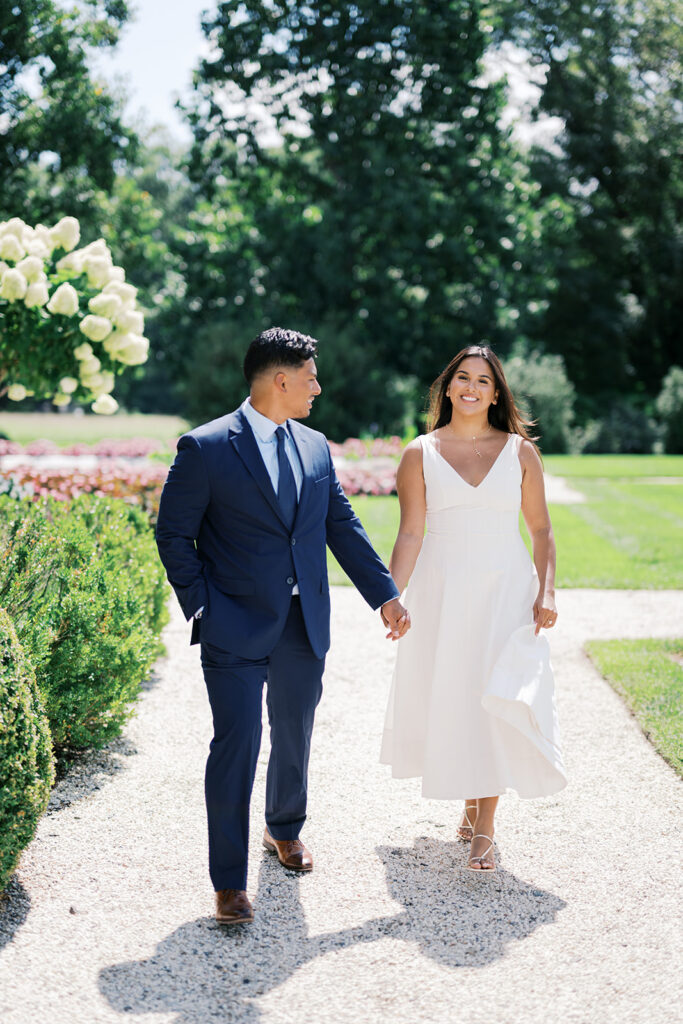 Nemours Estate engagement photos couple walking hand in hand on gravel path with hydrangeas white midi dress and navy suit summer Delaware