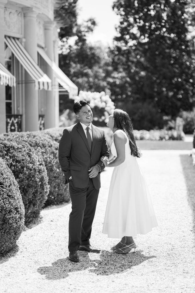 Black and white engagement photos candid laughing couple on gravel path beside Nemours Estate mansion facade with striped awnings Wilmington Delaware