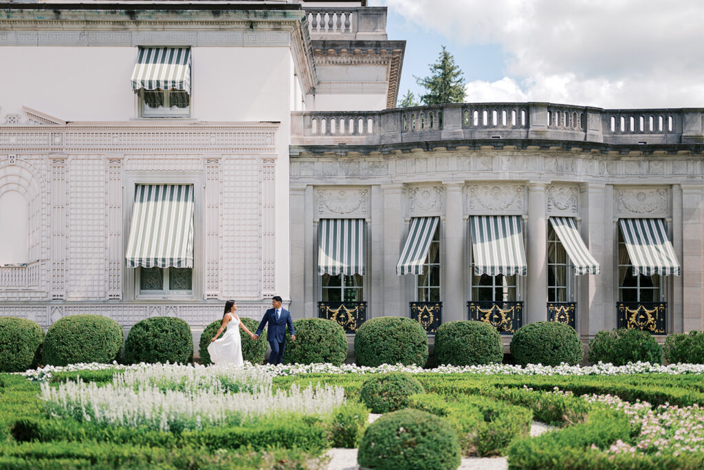 Nemours Estate engagement photography couple walking hand in hand along Nemours mansion facade with formal boxwood garden white dress and navy suit Wilmington Delaware